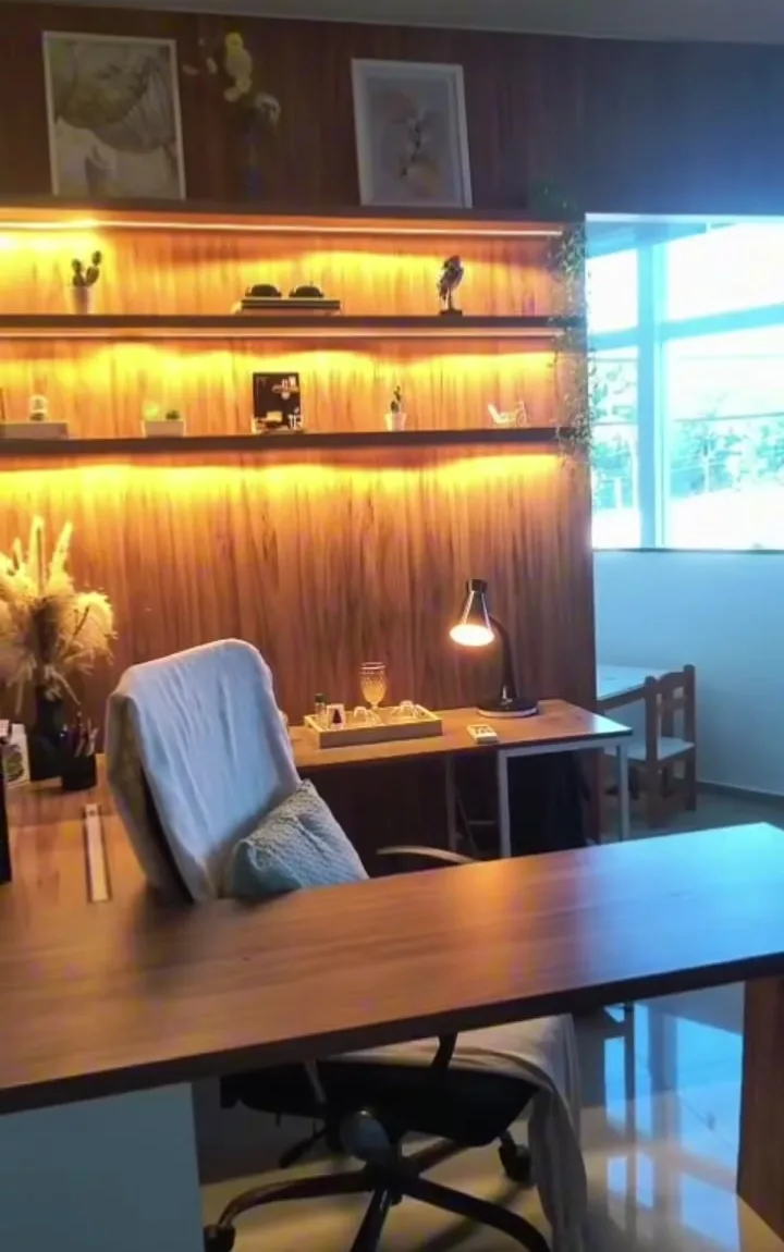 Mesa de atendimento do consultório, com estante de madeira iluminada ao fundo